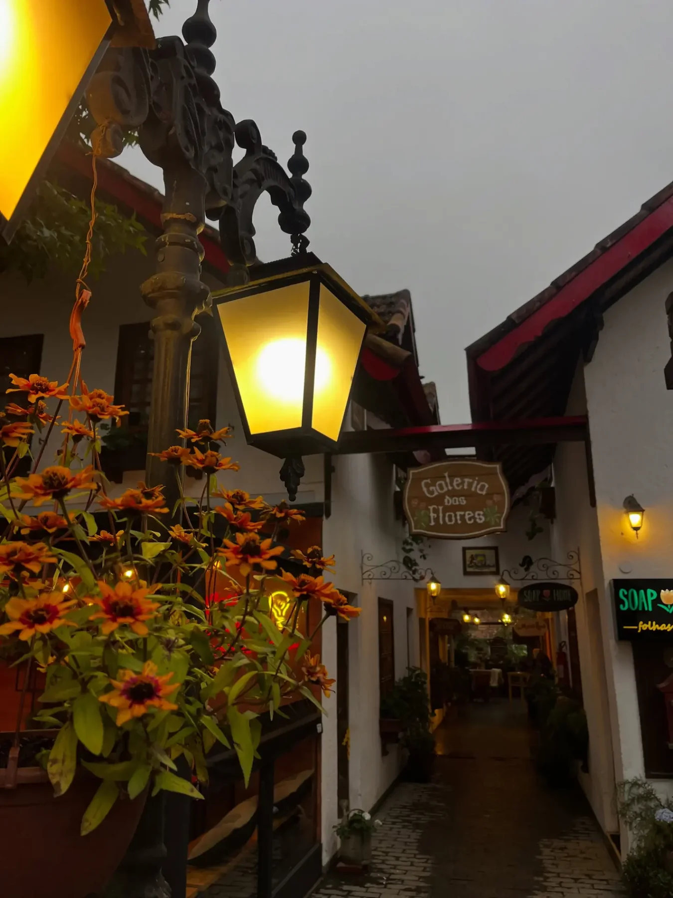 Galeria das Flores em Monte Verde MG ao entardecer, com luminária acesa e flores laranjas na entrada — passeio charmoso para dias de chuva